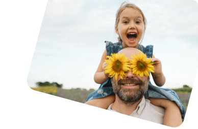 Ein Mann mit Bart trägt ein kleines Mädchen auf den Schultern. Seine Augen sind von Sonnenblumen verdeckt, das Mädchen lacht. 