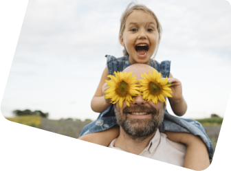 Ein Mann mit Bart trägt ein kleines Mädchen auf den Schultern. Seine Augen sind von Sonnenblumen verdeckt, das Mädchen lacht. 