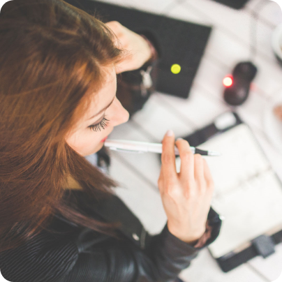 Eine junge Frau sitzt nachdenklich am Schreibtisch mit einem Stift in der Hand. Vor ihr liegen ein Notizblock, eine Computermaus und eine Tastatur. 