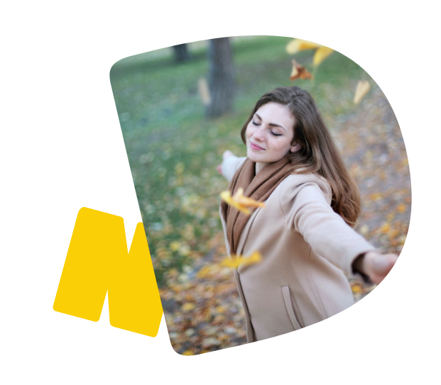 Eine junge Frau im Mantel steht mit geschlossenen Augen und ausgebreiteten Armen im Freien. Gelbe Blätter fallen auf sie nieder. 