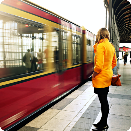Eine Frau in einem gelben Regenmantel steht an einem Bahnhof während ein Zug einfährt. 