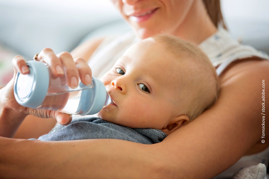 Ein junge trinkt Wasser aus einer Nuckelflasche. Er liegt dabei im Arm seiner Mutter und schaut in die Kamera. 