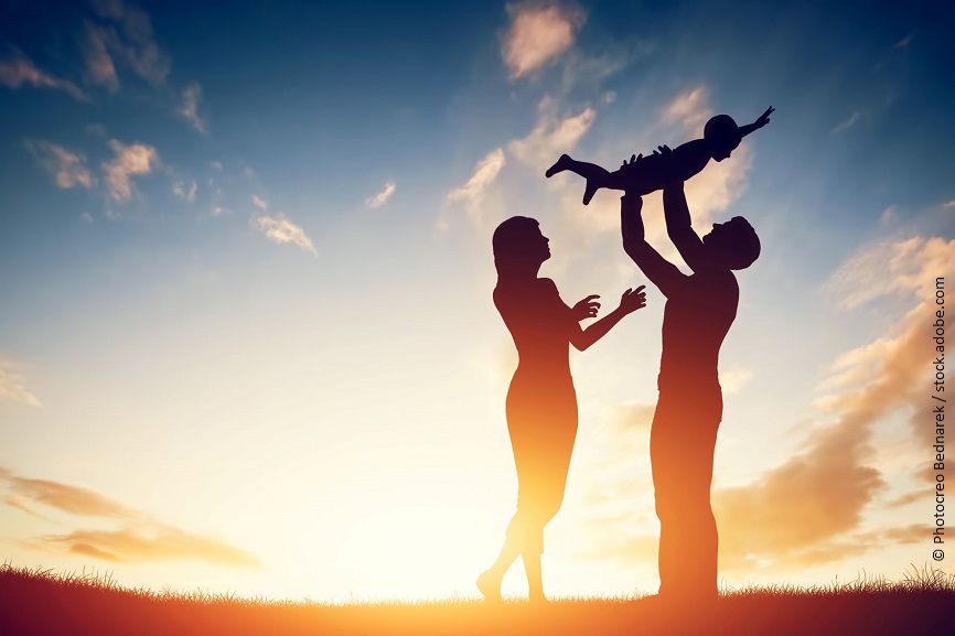 Eine dreiköpfige Familie steht vor einem Sonnenuntergang. Der Mann hält das Kind in höhe. 