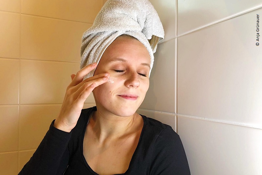 Anja steht mit geschlossenen Augen in ihrem Badezimmer und cremt sich das Gesicht ein. Ihre Haare sind in eine Handtuch gewickelt. 