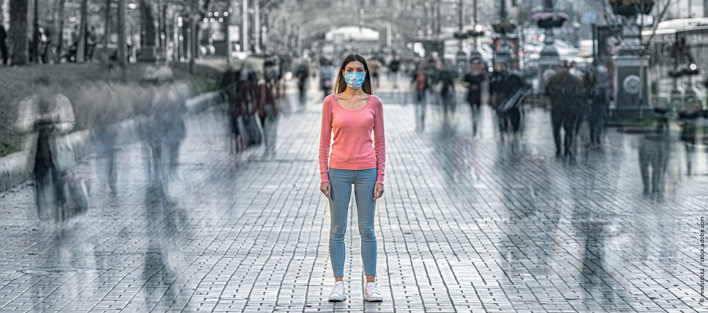 Eine Frau mit Maske steht auf der Straße. Links und Rechts gehen Menschen an ihr vorbei. 