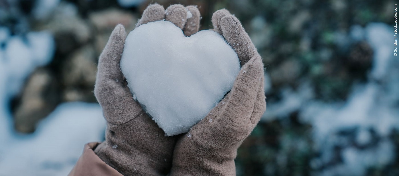 Hände in Handschuhen halten ein Herz aus Schnee. 