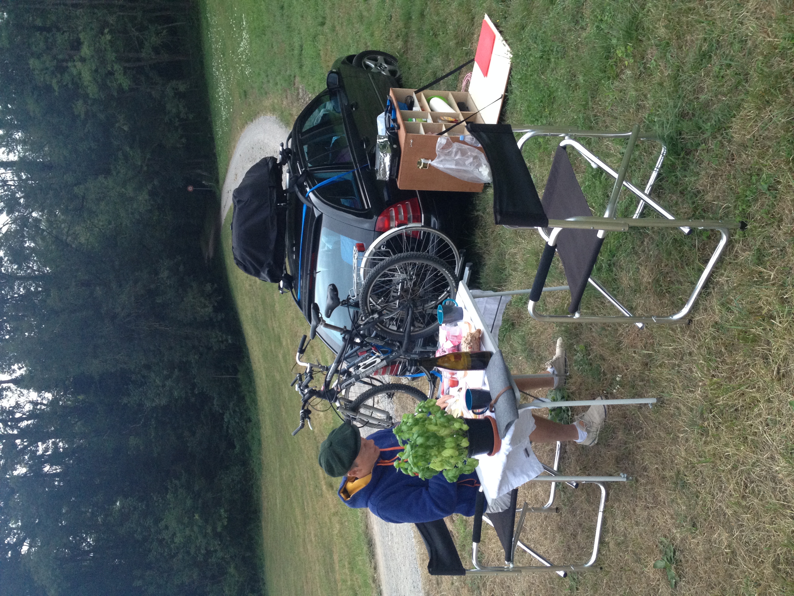 Ein Mann sitzt einen Campingtisch mit zwei Campingstühlen. Im Hintergrund parkt ein Auto mit zwei Fahrrädern. 