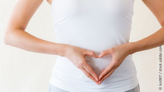 Eine Frau im weißen Top formt mit den Händen ein Herz vor ihrem Bauch.