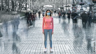 Eine Frau mit Maske steht in der Mitte der Straße, rechts und linkt gehen Menschen an ihr vorbei.  