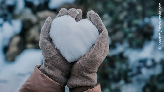 Behandschuhte Hände halten ein Herz aus Schnee in die Höhe. 
