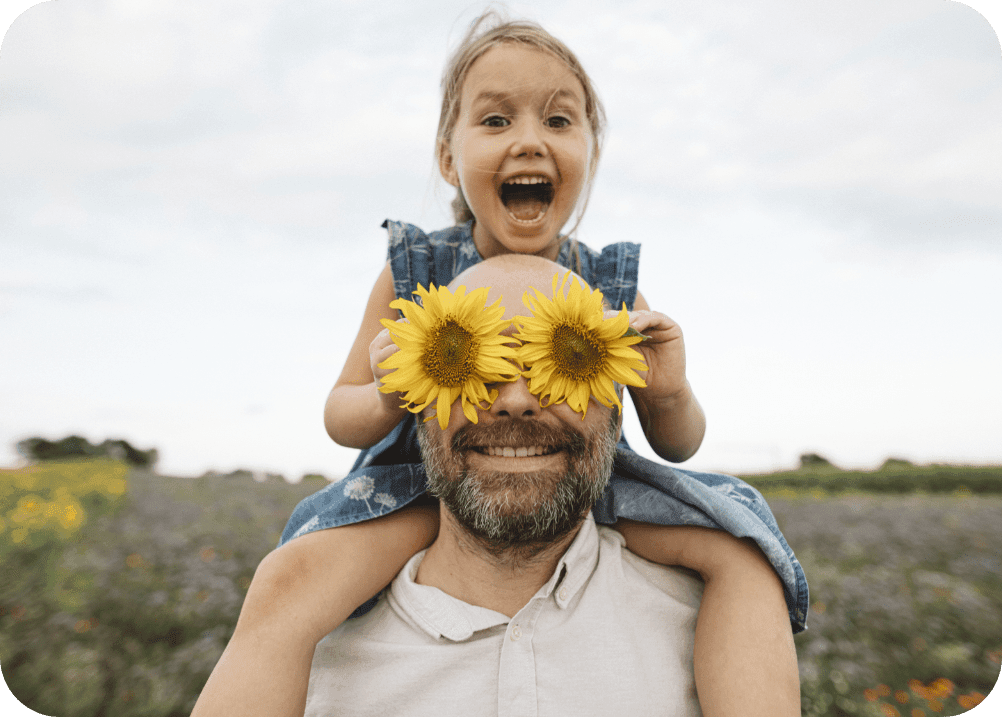 Ein Mann mit Bart trägt ein kleines Mädchen auf den Schultern. Seine Augen sind von Sonnenblumen verdeckt, das Mädchen lacht.