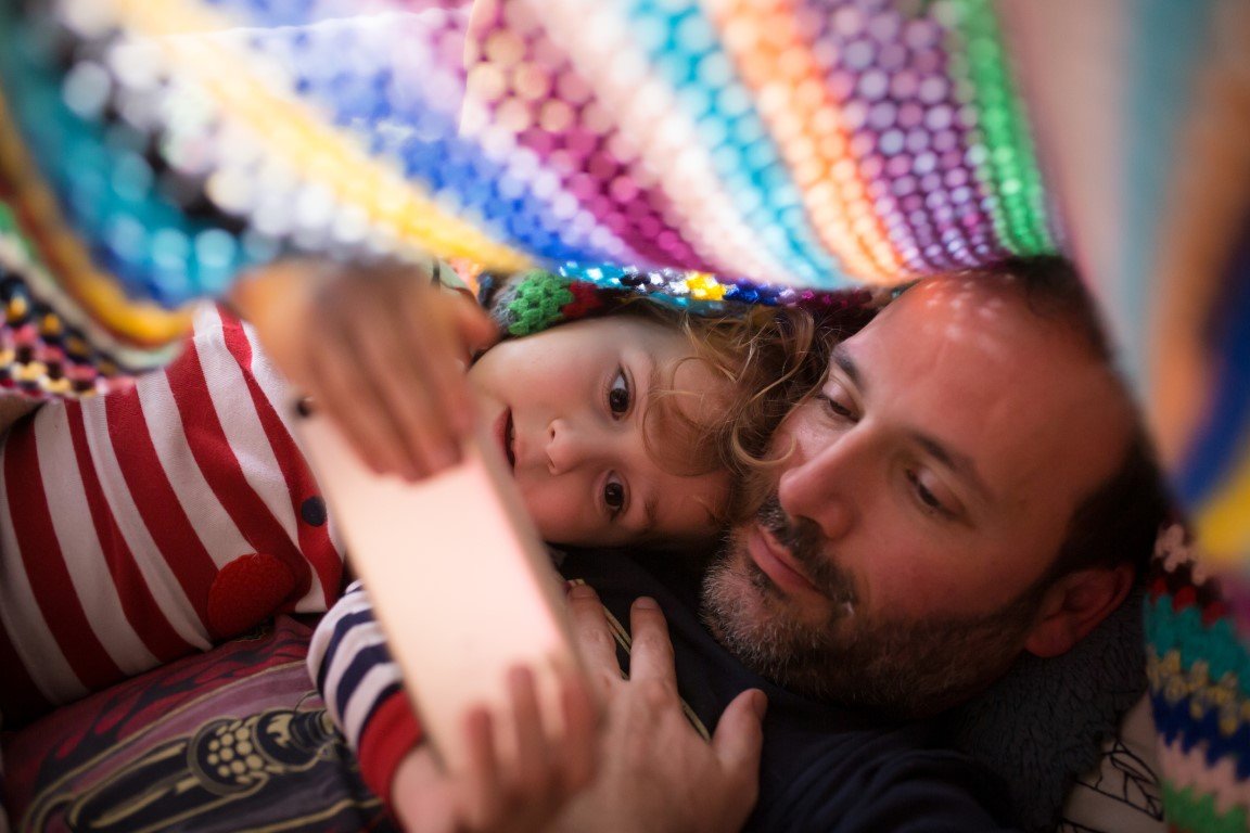 Ein Vater kuschelt mit seinem Sohn unter einer bunten Decke und sie schauen zusammen auf ein Handy.