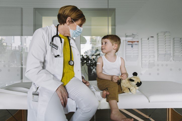 Eine Ärztin mit Maske spricht mit einem kleinen Jungen mit Teddaybär