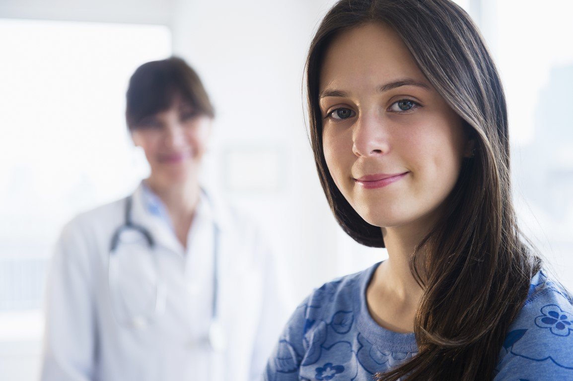 Ein junges Mädchen lächelt in die Kamera, im Hintergrund ist eine Ärztin zu sehen.