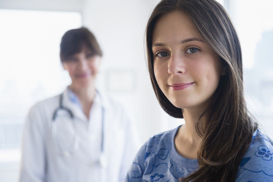Eine junge Patientin lächelt in die Kamera, im Hintergrund steht eine Ärztin.