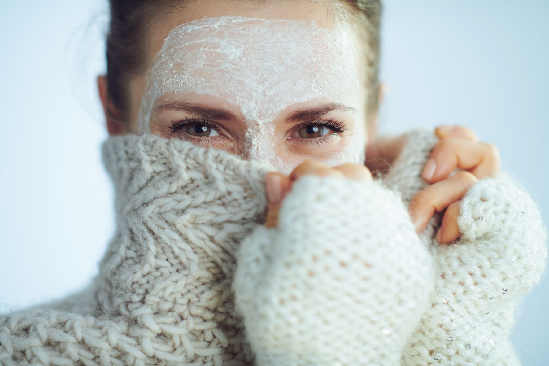 Eine Frau mit Creme im Gesicht versteckt dieses hinter einem Rollkragenpullover.